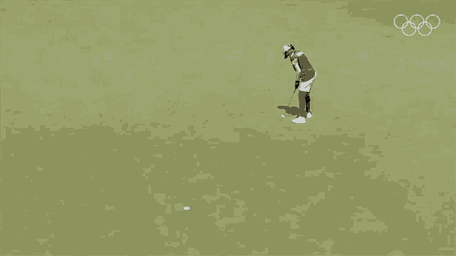 a woman is putting a golf ball on a green with the olympic rings visible in the background