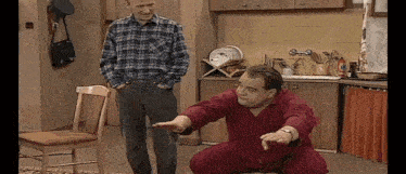a man squatting down in a kitchen while another man stands in the background .