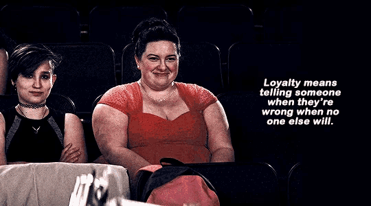 a woman in a red dress sits next to a woman in a black dress in a dark auditorium