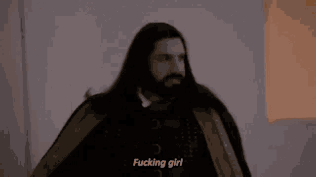 a man with long hair and a beard is standing in front of a wall and says `` fucking girl '' .
