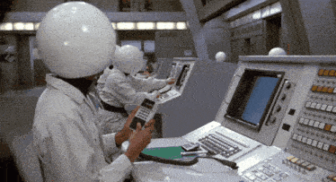 a man wearing a white helmet sits at a control panel holding a phone