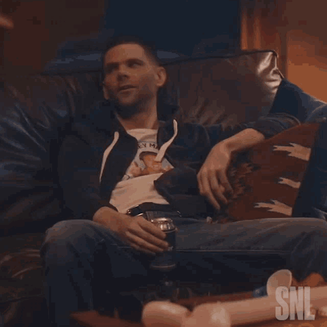 a man is sitting on a couch with a can of beer and a snl logo behind him