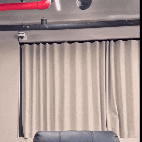 a couch sits in front of a window with white curtains and a red pipe hanging from the ceiling