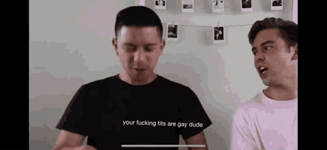 a man wearing a black shirt that says your fucking tits are gay dude talks to another man in a pink shirt .