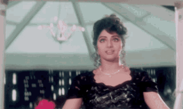 a woman in a black dress and necklace is standing in a room with a chandelier hanging from the ceiling .