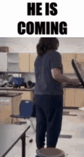 a man is standing on a stool in a kitchen with the words `` he is coming '' .