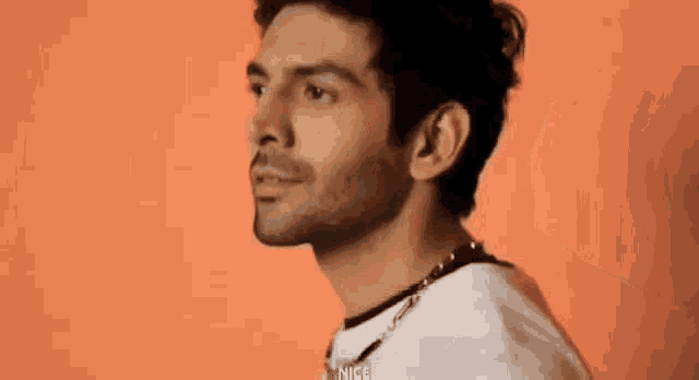 a man wearing a white t-shirt and a necklace is smiling against an orange background .