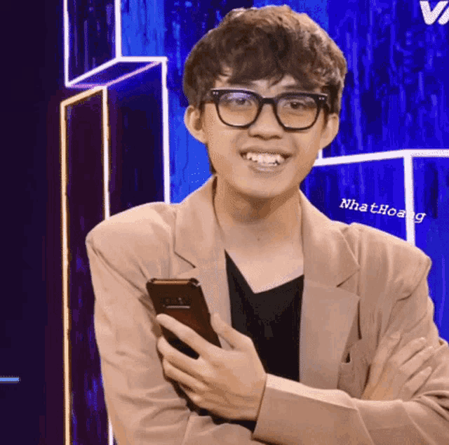 a man wearing glasses and a tan jacket holds a cell phone
