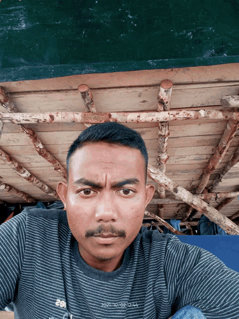 a man with a mustache is taking a selfie in front of a wooden structure with a date stamp of 2021/10/02 12:44