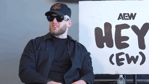 a man wearing sunglasses sits in front of a sign that says hey