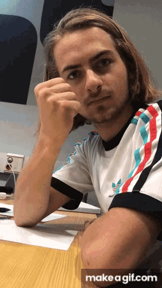 a man with long hair is sitting at a table with his hand on his chin and a make a gif.com button below him