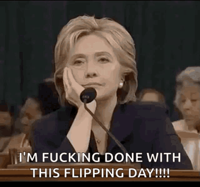 hillary clinton is sitting at a podium with her hand on her chin and a microphone in front of her .