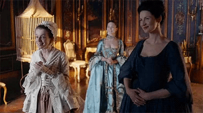 three women in historical costumes are standing in a room with a bird cage in the background .