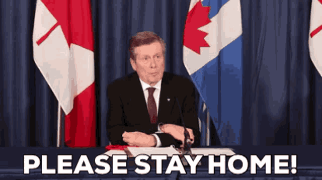 a man in a suit and tie is sitting at a table with canadian flags behind him and says please stay home