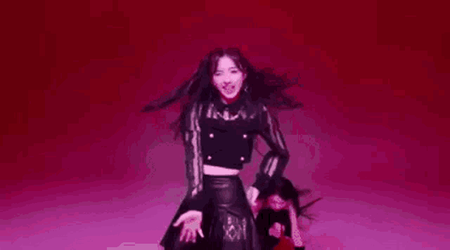 a woman in a black crop top and leather skirt is dancing in front of a pink background .