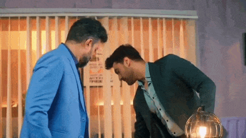 two men in suits are standing next to each other in front of a window and talking to each other .