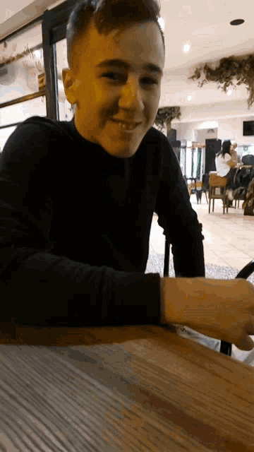 a young man in a black sweater sits at a wooden table