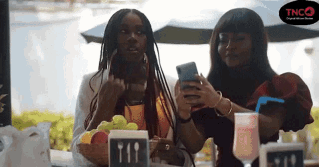 two women are sitting at a table looking at their cell phones with a tnco logo behind them