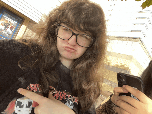 a girl wearing glasses and a shirt that says ' apocalypse ' on it points at her phone