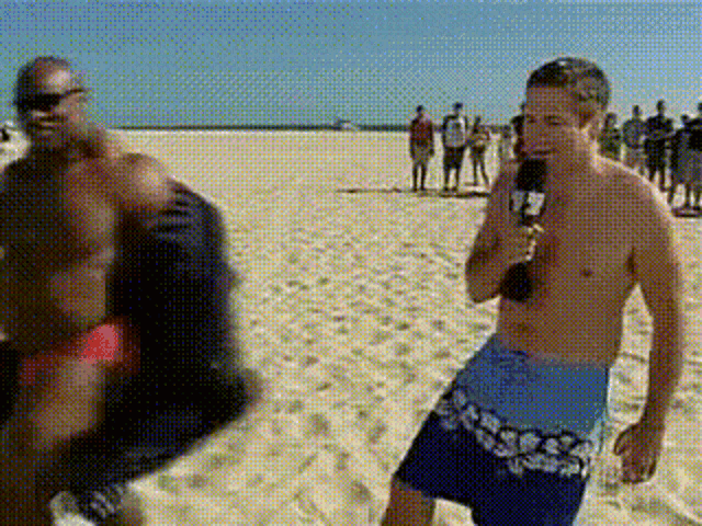 a shirtless man holding a microphone stands on a beach next to another man