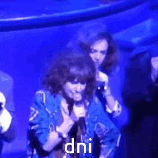 a woman in a blue jacket is singing into a microphone and the word dni is on the screen behind her .
