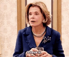a woman in a blue suit is holding a glass of wine and saying `` prove it '' .