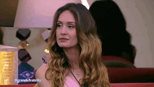 a woman in a pink dress is sitting on a red couch in front of a lamp with the hashtag grandefratello on the bottom