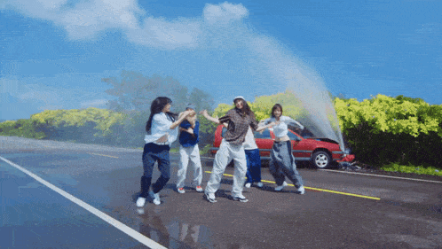 a group of people are dancing on a road in front of a red car