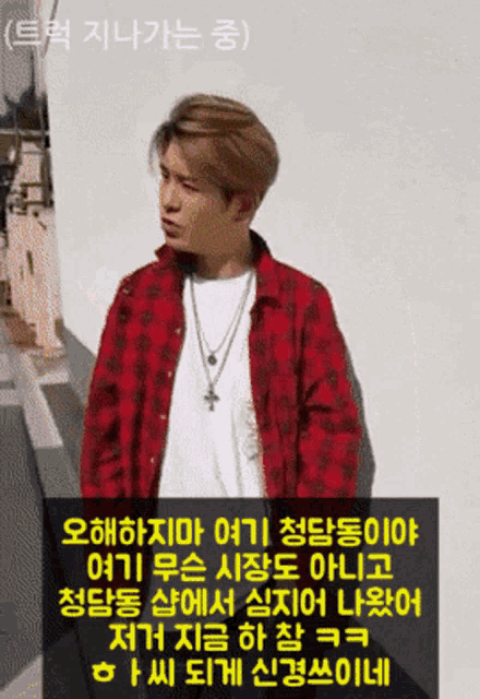 a man wearing a red plaid shirt and a cross necklace stands in front of a white wall