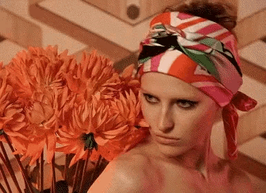 a woman wearing a colorful head scarf stands next to a vase of orange flowers