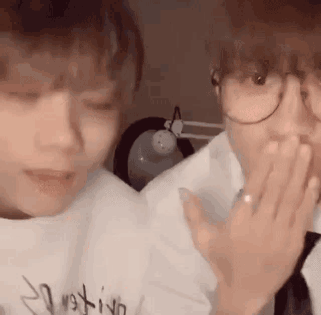 two young men wearing glasses are sitting next to each other and one of them is covering his mouth with his hand .