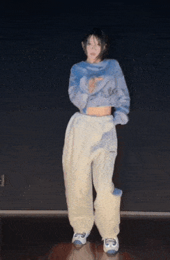 a woman in a blue sweater and white pants is dancing in a dark room .