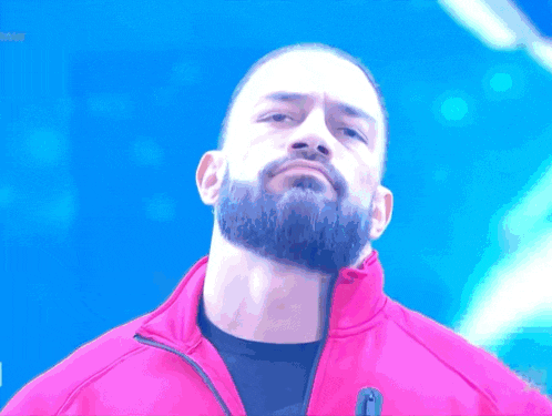 a man with a beard wearing a pink jacket stands in front of a blue background