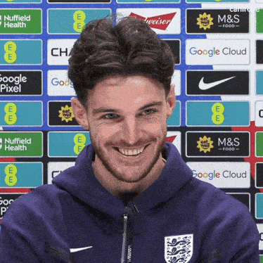 a man wearing a purple hoodie is smiling in front of a wall of advertisements including m & s food and google cloud