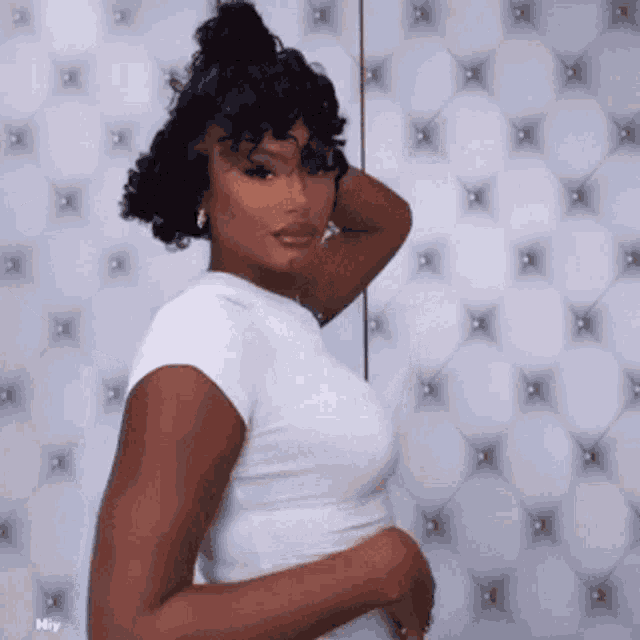 a woman with curly hair is wearing a white shirt and standing in front of a white quilted wall .