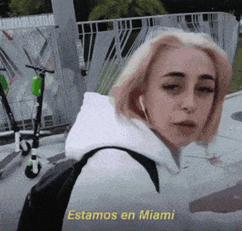 a girl in a white hoodie is standing in front of scooters and says " estamos en miami "