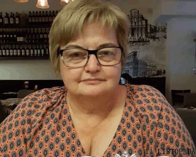 a woman wearing glasses sits in front of a wall with wine bottles on it