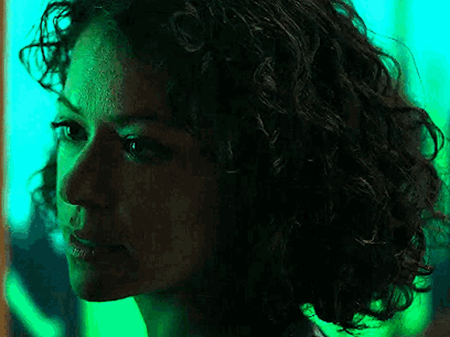 a woman with curly hair is looking at the camera in a dark room with green lights behind her .