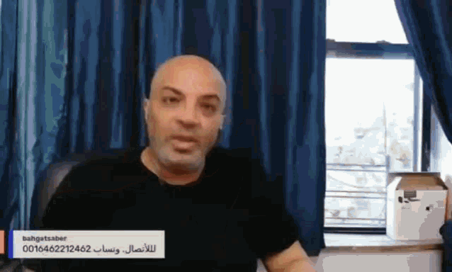 a bald man with a beard is sitting in front of a window with a blue curtain ..