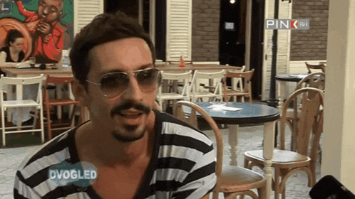 a man wearing sunglasses and a striped shirt is sitting in front of tables and chairs with a pink sign above him