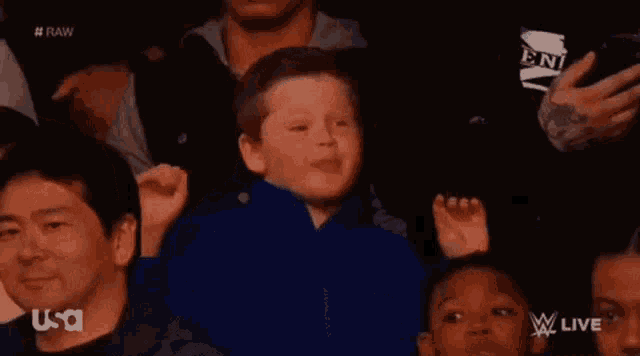 a young boy in a blue jacket is sitting in a crowd of people watching a wrestling match .