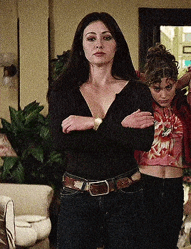 two women are standing in a living room with their arms crossed . one of the women is wearing a crop top .