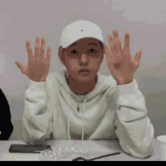 a person wearing a white hat and glasses is sitting at a table with their hands up .