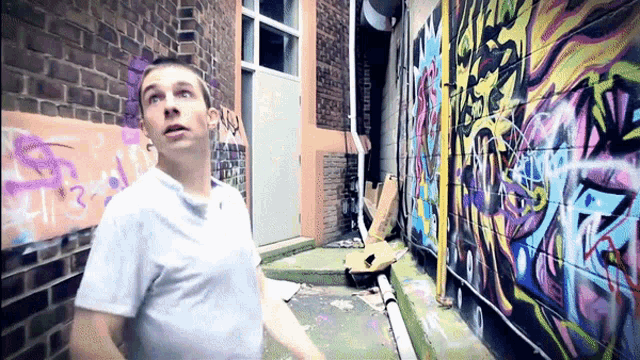a man in a white shirt stands in a narrow alleyway with graffiti on the walls