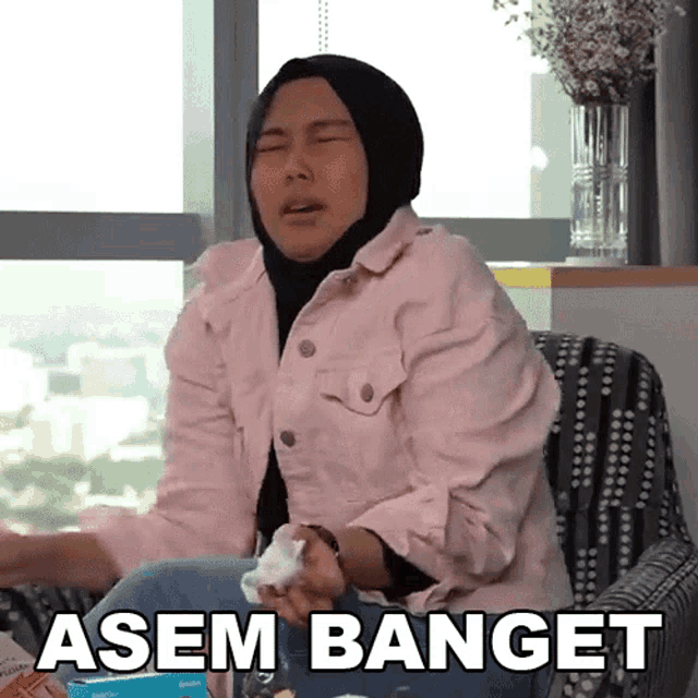 a woman wearing a hijab and a pink jacket is sitting in a chair with the words asem banget written below her