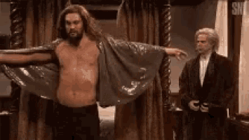 a shirtless man is standing in front of a bed with his arms outstretched while another man stands behind him .