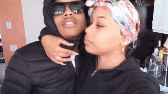 a man wearing a hooded jacket and sunglasses kisses a woman wearing a head scarf