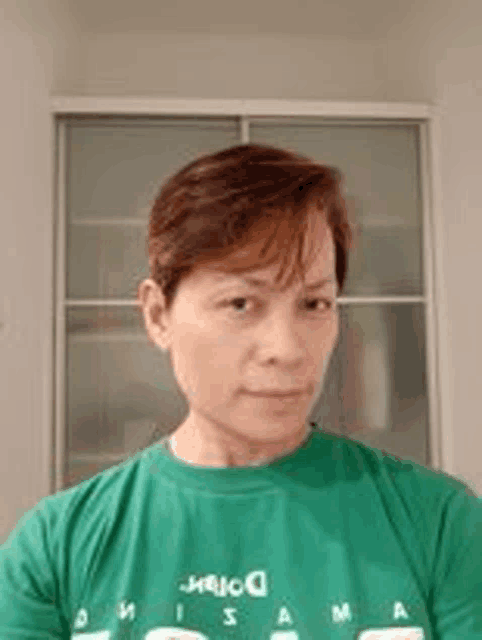 a woman wearing a green t-shirt is taking a selfie in front of a window .