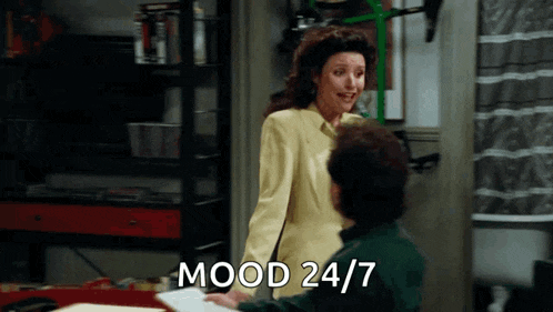 a woman in a yellow suit is standing next to a man in a green shirt and says mood 24/7 .