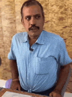 a man with a mustache wearing a blue shirt is sitting at a table with his hands on his hips .